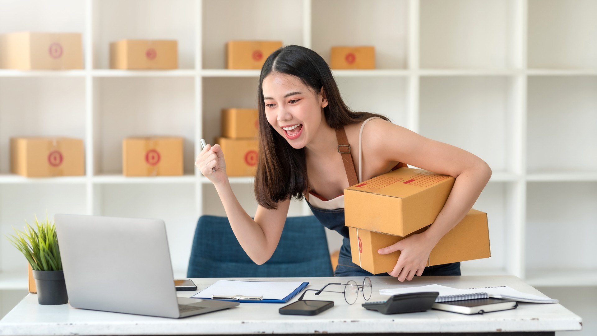 Asian small business owner wearing an apron is successful selling online using laptop computer. Happy Asian Business Woman Successful Excited Raised Hands Rejoicing.